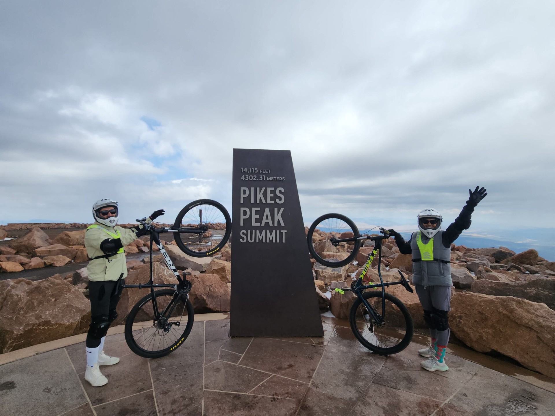 Pikes Peak Cog Up · Bike Down Two cyclists in helmets and pads celebrating with their mountain bikes at the 14,115 ft Pikes Peak Summit marker